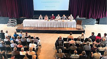 Podium mit sechs Personen bei einer Veranstaltung vor Publikum in einem Saal mit rotem Vorhang und Projektionsleinwand auf der steht "Energiegenossenschaft Schwerte eG, Schwerte 23.06.2025"