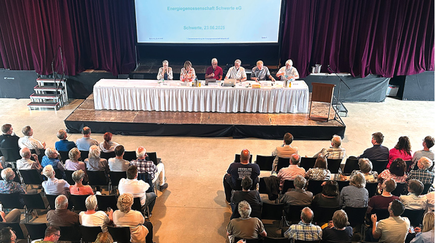 Podium mit sechs Personen bei einer Veranstaltung vor Publikum in einem Saal mit rotem Vorhang und Projektionsleinwand auf der steht "Energiegenossenschaft Schwerte eG, Schwerte 23.06.2025"